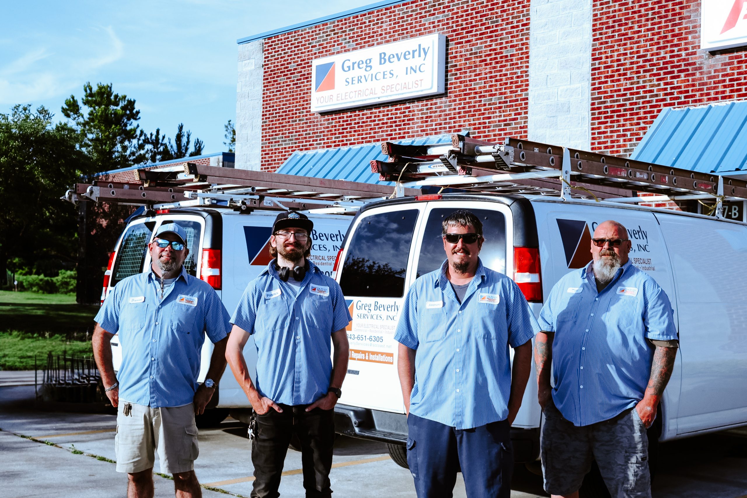 Four Greg Beverly Services electricians standing in front of branded service vans outside the company headquarters. The team members wear company uniforms, with ladder-equipped electrical service vehicles parked behind them.