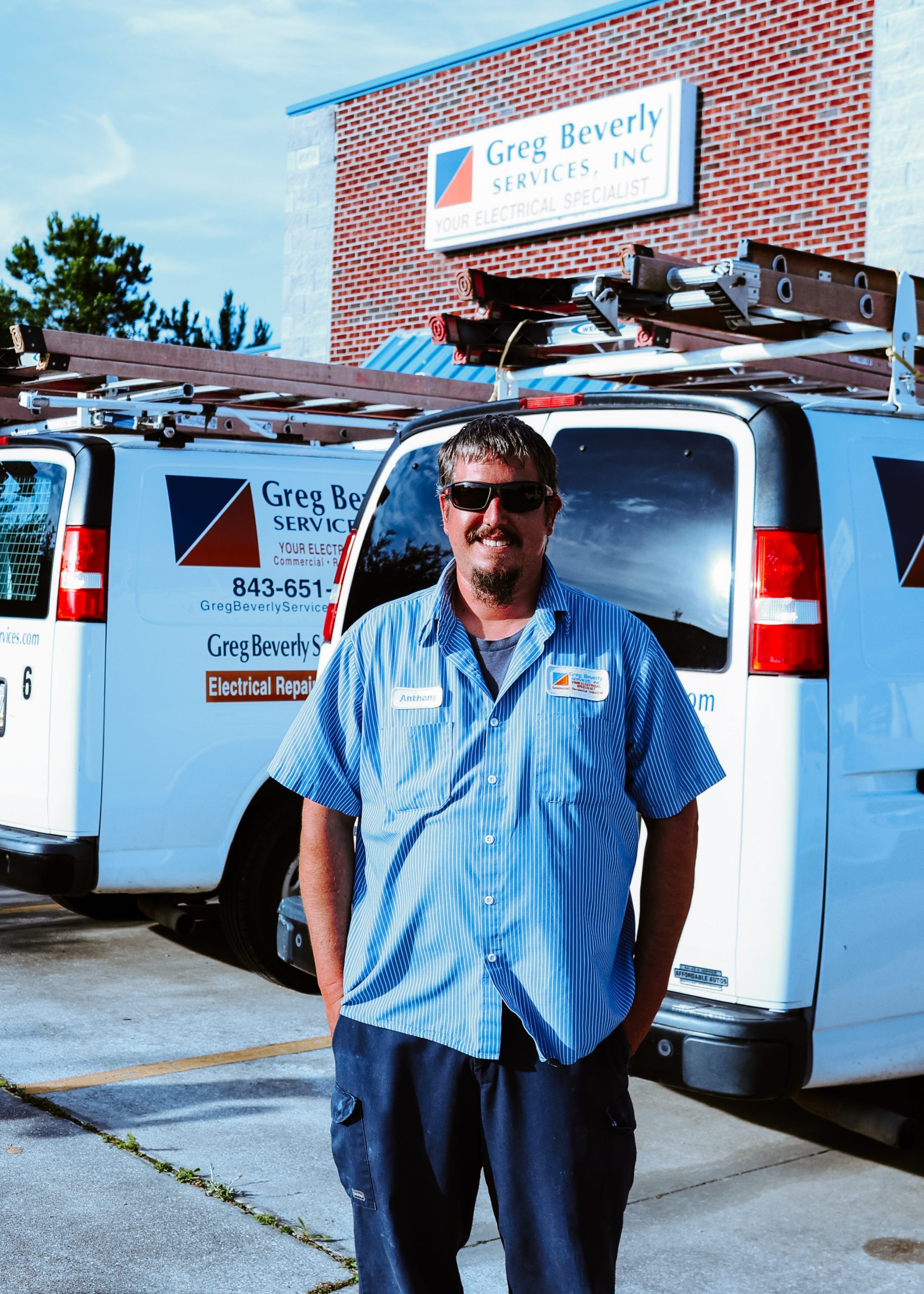 Greg Beverly Services electrician standing in front of company service vans outside the company headquarters. The technician wears a branded work uniform, with ladder-equipped electrical service vehicles parked behind him.