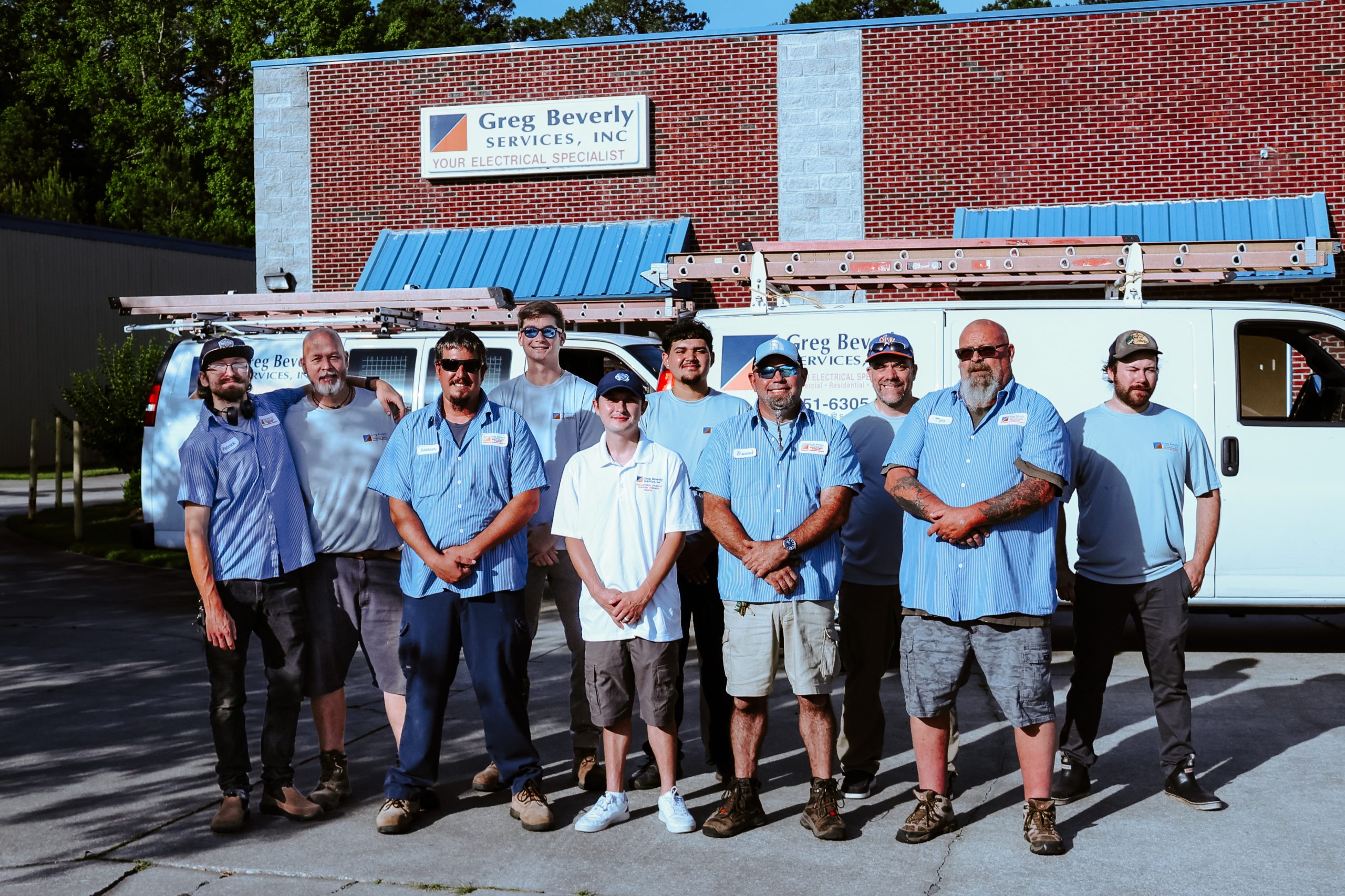 Team photo of Greg Beverly Services electricians standing in front of company service vans and the Greg Beverly Services office building. The crew wears branded work uniforms and poses outside the electrical contractor’s headquarters on a sunny day.